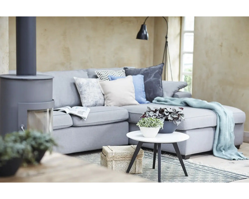 Moderne woonkamer met grijze hoekbank, sierkussens, lichtblauwe plaid, witte salontafel met planten en een grijze houtkachel.
