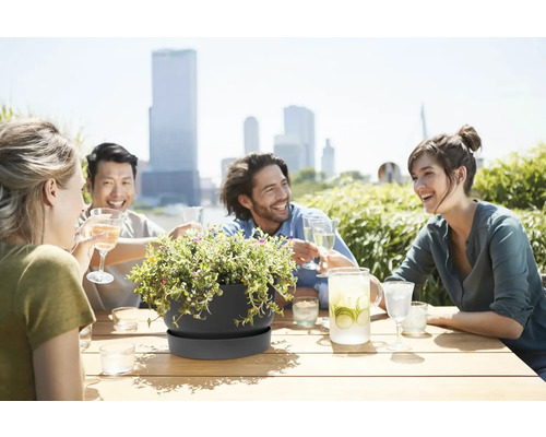 Donkergrijze plantenschaal met schotel op houten tafel, op de achtergrond vrienden op een zonnig dakterras met skyline.