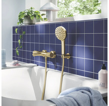 Gouden badmengkraan met handdouche op blauwe tegelwand boven wit bad, lichte badkamer met plant.