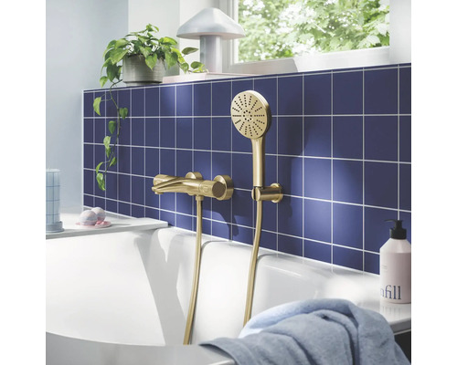 Gouden badmengkraan met handdouche op blauwe tegelwand boven wit bad, lichte badkamer met plant.