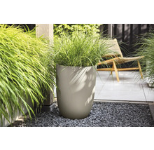 Hoge beige plantenbak met golvende structuur en siergras op donker grind in een lichte tuin met een terras en houten paal.