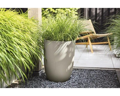 Hoge beige plantenbak met golvende structuur en siergras op donker grind in een lichte tuin met een terras en houten paal.