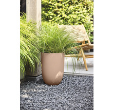 Hoge ronde plantenbak met een golvende textuur en siergras op een grindondergrond in een tuin naast een houten paal en een tuinstoel.