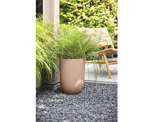 Hoge ronde plantenbak met een golvende textuur en siergras op een grindondergrond in een tuin naast een houten paal en een tuinstoel.