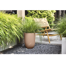 Bruine ronde bloempot met geribbelde structuur en groen siergras op een terras met donker grind naast een houten stoel.