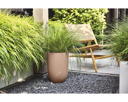 Bruine ronde bloempot met geribbelde structuur en groen siergras op een terras met donker grind naast een houten stoel.