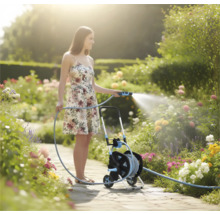 Vrouw besproeit een bloeiende tuin met een tuinslang op een slangenwagen op een licht tuinpad.