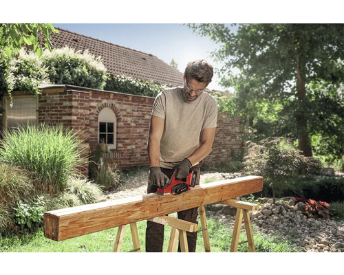 Een man gebruikt een elektrische schaaf op een houten balk op schragen in een lichte tuin. Hij draagt een veiligheidsbril en handschoenen.