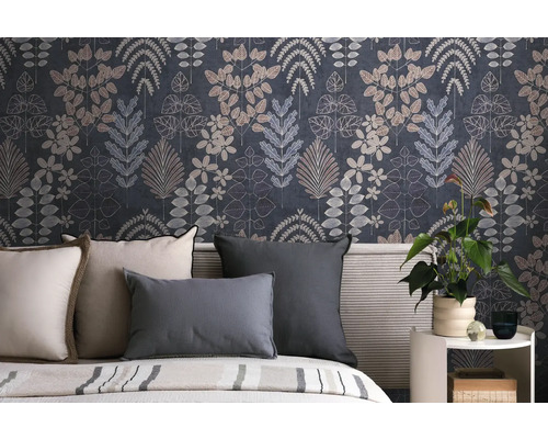 Vliesbehang met botanisch bladerpatroon in donkerblauw en grijs achter een bed met kussens en een kamerplant op een bijzettafel.