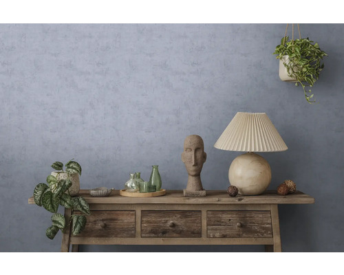 Rustiek houten dressoir voor een blauwgrijze muur met textuur, voorzien van een tafellamp, stenen sculptuur, glazen flessen en kamerplanten.