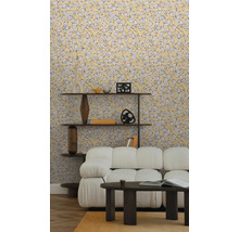 Vliesbehang met geel bladmotief in een lichte woonkamer met witte bank, donkere salontafel en houten wandrek op parketvloer.