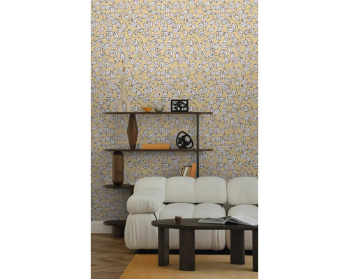 Vliesbehang met geel bladmotief in een lichte woonkamer met witte bank, donkere salontafel en houten wandrek op parketvloer.