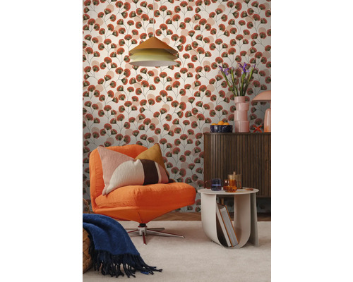 Woonkamer met patroonbehang met gestileerde terracotta en groene bladeren. Met oranje corduroy fauteuil, een houten kast en een bijzettafel.