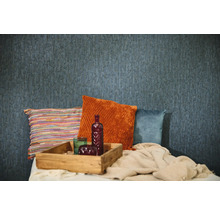 Blauw behang met structuur in een kamer met decoratieve kussens in oranje, blauw en meerkleurig, een houten dienblad en een deken.