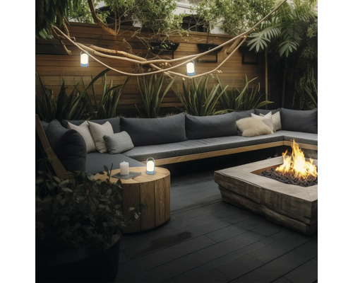 Modern terras met L-bank, kussens, vuurplaats, houten tafel en draagbare LED-lampen tussen planten en een houten wand.