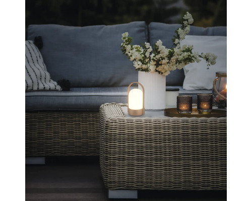 Draagbare LED-lantaarn op een gevlochten salontafel in een tuinlounge met bank, kussens en een bloemenvaas.