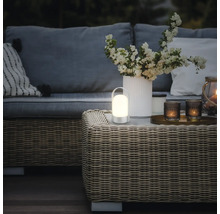 Draagbare, warm verlichte LED-tafellamp op een rotan tafel in een modern ingerichte tuinlounge met bank en bloemen.