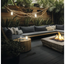 Moderne tuinlounge met een grijze bank, kussens, een vuurplaats, een houten bijzettafel met lantaarn en sfeervolle buitenverlichting.