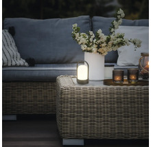 Draagbare LED-tafellamp voor buiten op een rotan tafel in een warm verlichte tuinlounge met bank, kussens en een vaas met bloemen.