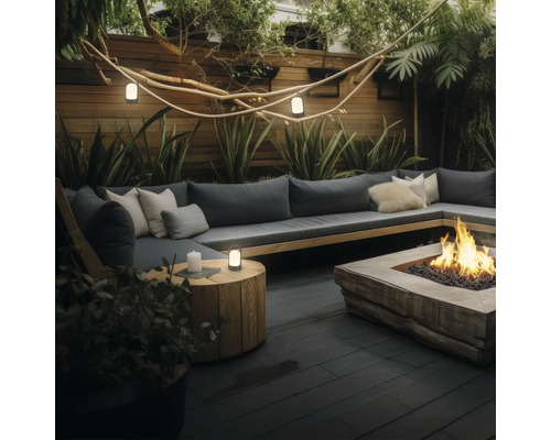 Moderne buitenlounge met grote L-vormige houten bank, donkergrijze kussens en een centrale vuurplaats op een donker terras omgeven door planten