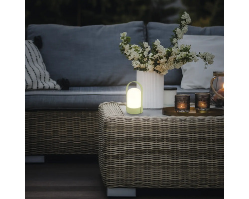 Draagbare LED-tafellamp op een gevlochten tuintafel in een sfeervol verlichte buitenruimte met bank en bloemen.