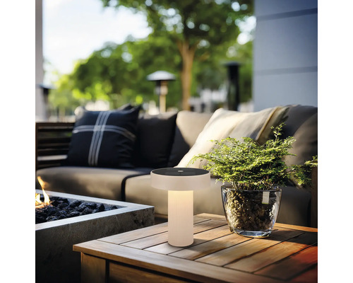 Moderne solar tafellamp voor buiten met warm licht op een houten tafel in een loungehoek met bank en plant.