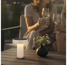 Moderne solar tafellamp op een houten terrastafel in een tuin bij avondschemering met bloemen en mensen op de achtergrond.