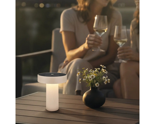 Moderne solar tafellamp op een houten terrastafel in een tuin bij avondschemering met bloemen en mensen op de achtergrond.