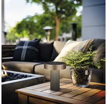 Moderne tafellamp op een houten tafel op een licht tuinterras met bank, vuurplaats en plant in glazen pot.