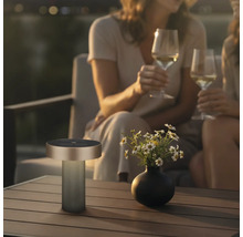 Moderne draagbare outdoor tafellamp op een houten tafel naast een bloemenvaas in een gezellige avondsetting met mensen op de achtergrond.