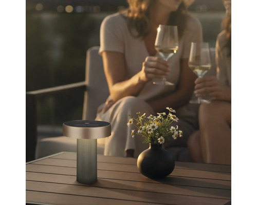 Moderne draagbare outdoor tafellamp op een houten tafel naast een bloemenvaas in een gezellige avondsetting met mensen op de achtergrond.