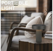 Moderne draagbare LED-tafellamp op een houten tafel in een buitenruimte met fauteuils.