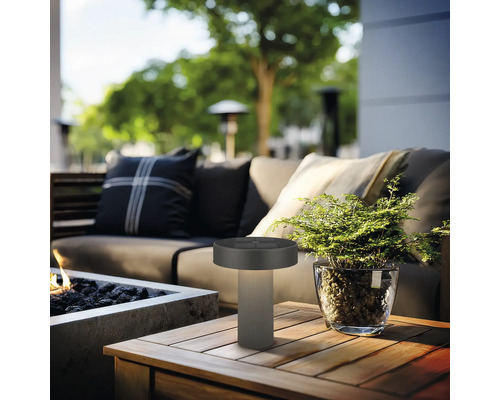 Moderne buiten-tafellamp op een houten tafel in een georganiseerde tuinlounge met bank, plant en vuurplaats.