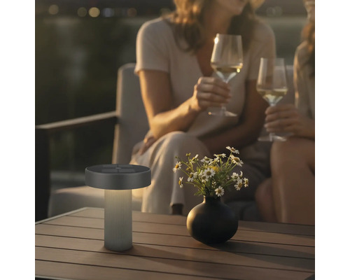 Moderne LED-tafellamp op zonne-energie voor buiten op een houten tafel in een gezellige avondtuin met mensen.