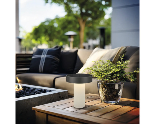 Moderne LED-tafellamp voor buiten op een houten tafel in een gezellige zithoek met vuurkorf en planten.