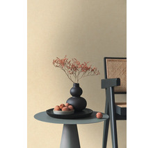 Beige vliesbehang met fijne textuur in een minimalistische ruimte met bijzettafel, zwarte vaas en rotan stoel.