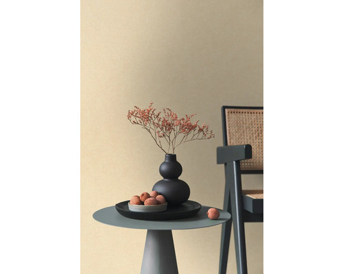 Beige vliesbehang met fijne textuur in een minimalistische ruimte met bijzettafel, zwarte vaas en rotan stoel.