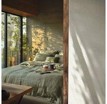 Lichte slaapkamer met bed in saliegroen linnen beddengoed, houten accenten en een groot raam met uitzicht op groen in zonlicht.