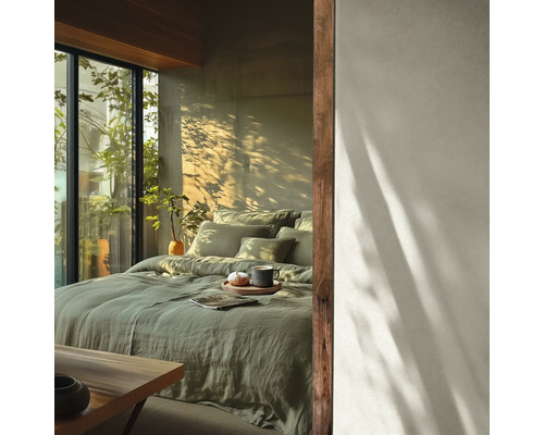 Lichte slaapkamer met bed in saliegroen linnen beddengoed, houten accenten en een groot raam met uitzicht op groen in zonlicht.