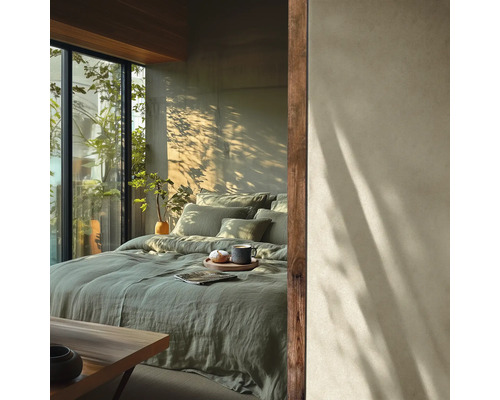 Lichte slaapkamer met saliegroen linnen beddengoed op een groot bed bij een betonnen muur met zonlicht en uitzicht op groen door grote ramen.