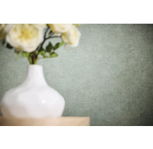 Effen vliesbehang met fijne linnenstructuur in een lichte groentint achter een witte vaas met bloemen.