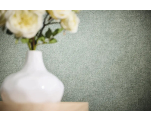 Effen vliesbehang met fijne linnenstructuur in een lichte groentint achter een witte vaas met bloemen.