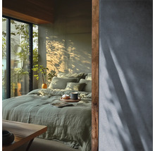 Lichte slaapkamer met groen linnen beddengoed op het bed, een gestructureerde muur en houten accenten naast een raam met uitzicht op groen.