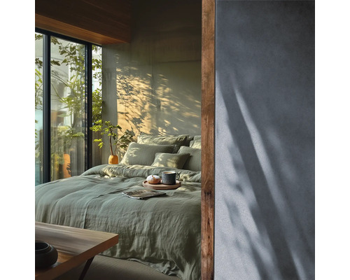 Lichte slaapkamer met groen linnen beddengoed op het bed, een gestructureerde muur en houten accenten naast een raam met uitzicht op groen.