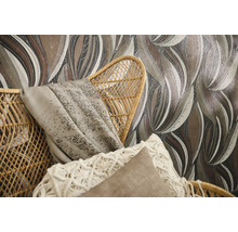 Vliesbehang met grootbladig patroon in aardetinten achter een gevlochten rotan stoel met kussens en plaid.