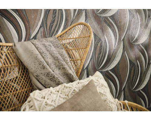 Vliesbehang met grootbladig patroon in aardetinten achter een gevlochten rotan stoel met kussens en plaid.