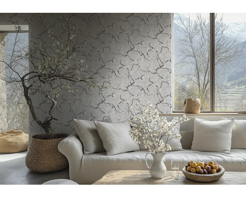 Vliesbehang met een fijn takken- en bloemenpatroon op een wand in een lichte woonkamer achter een bank met kussens.