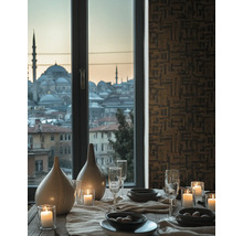 Gedekte eettafel met kaarsen, glazen en vazen voor een raam met stadszicht en behang met geometrisch patroon.