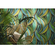 Vliesbehang met abstract bladerpatroon achter een rotan stoel met kussens en een kamerplant.