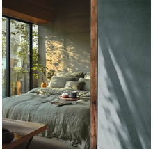 Moderne slaapkamer met groen linnen beddengoed, groot raam met uitzicht op groen en natuurlijk licht met schaduwen van planten op de muur.